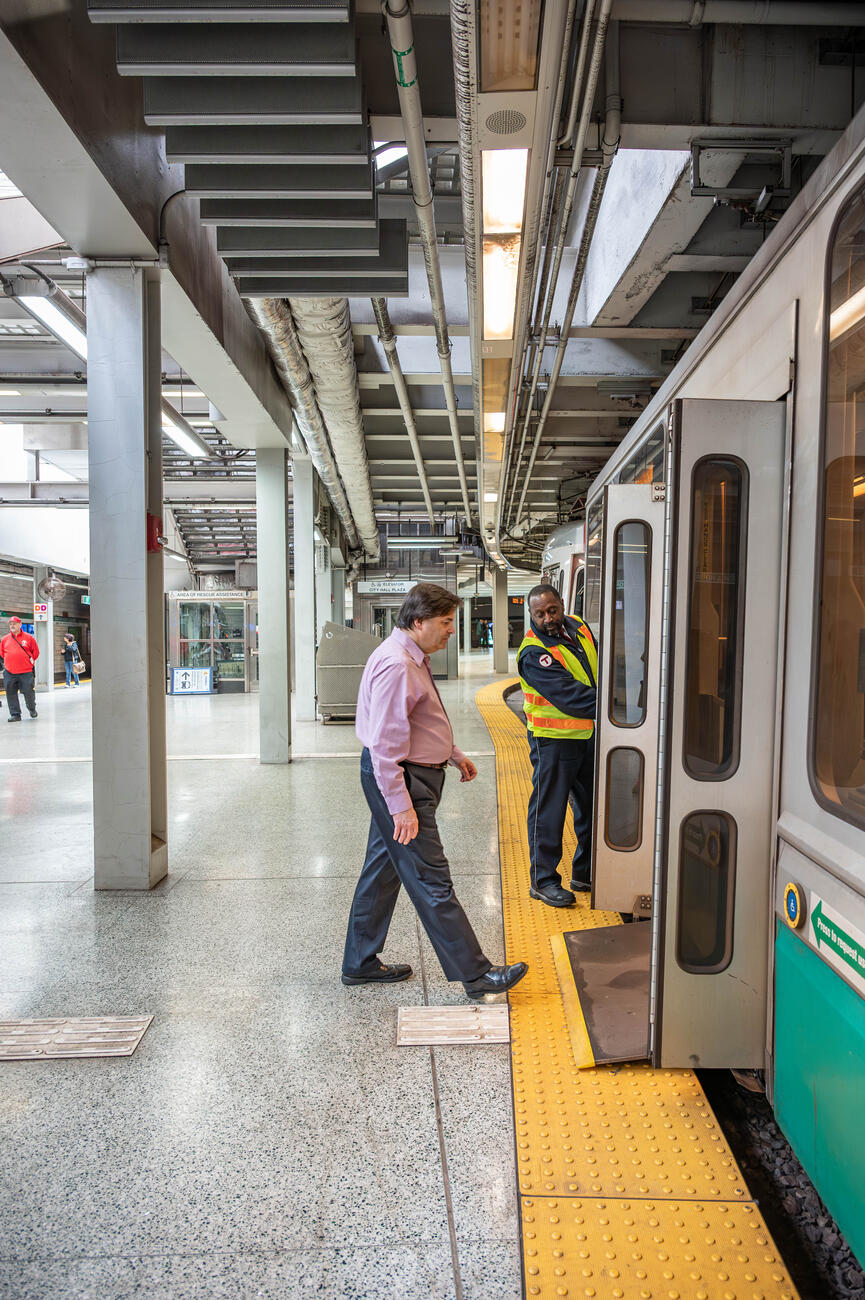 A green line inspector, an African American man, deploys the onboard ramp for a white male rider with a non-apparent disability.