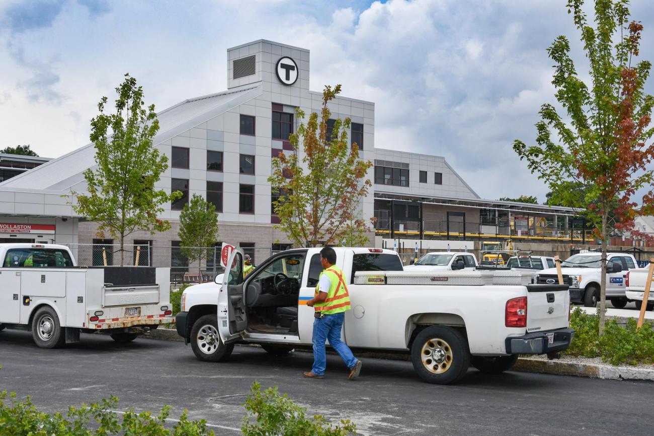 The facade of the renovated Wollaston Station in the background, with the parking lot in the forground. A construction crewperson is at his pick-up truck. (August 7, 2019)