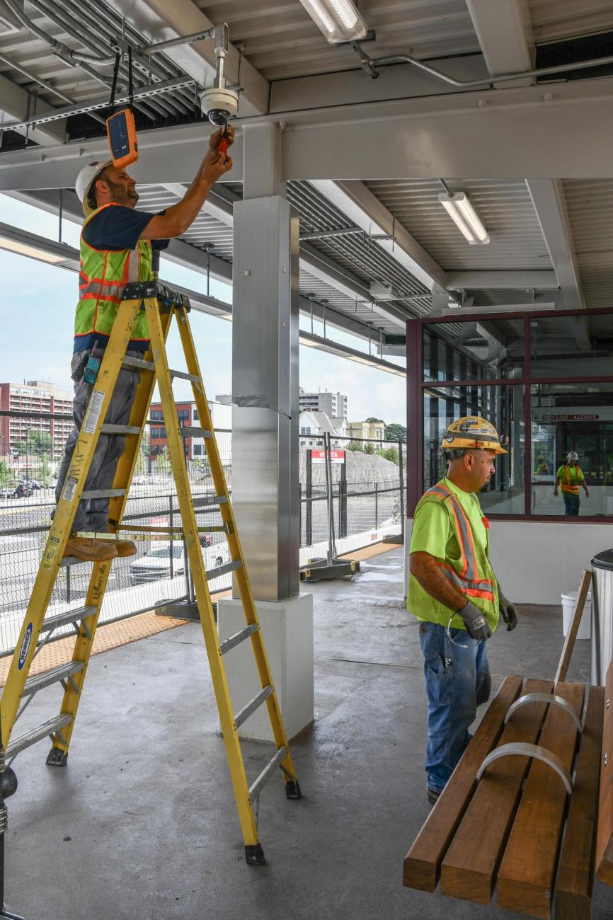 A crewperson on a ladder and another crewperson nearby at Wollaston Station, during the final stages of renovation. The parking lot is in the background. (August 7, 2019)