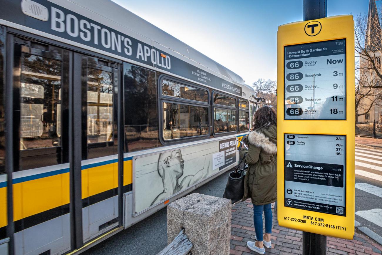 A rider gets ready to board a bus near Harvard Square as an E Ink sign displays real-time information
