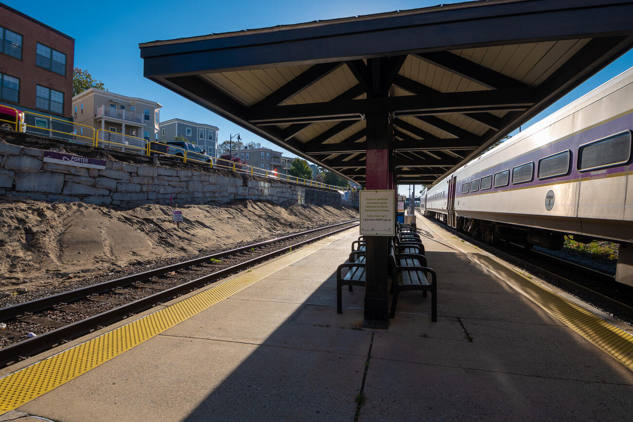 Empty Commuter Rail platform at Porter Square during a clear fall day. The Fitchburg train is seen going inbound, while the retaining wall under Somerville Ave is under construction.