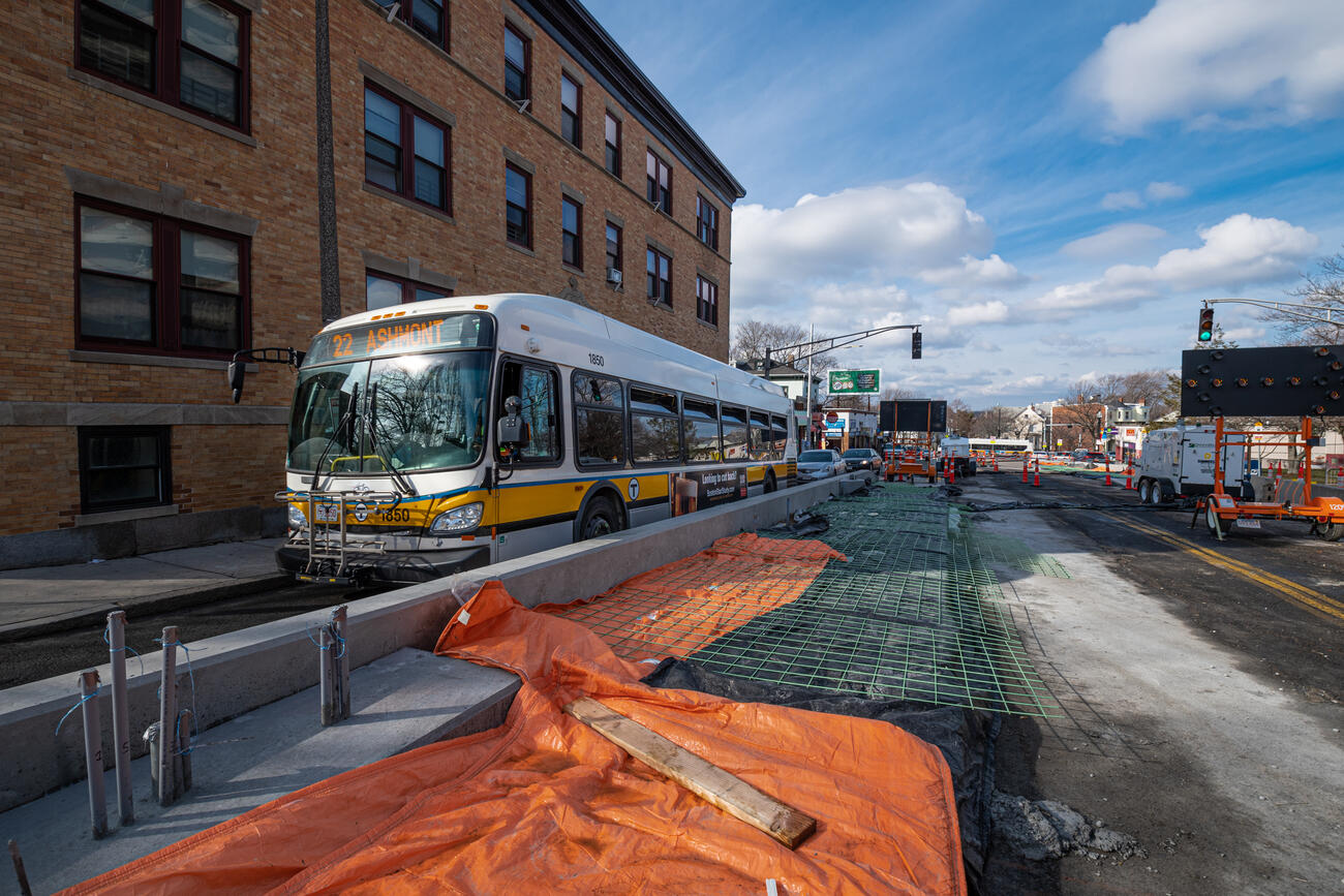 A Route 22 bus inbound to Ashmont passes by a new concrete bus boarding platform under construction near the intersection of Columbus Ave and Dixwell St near Egleston Square. The concrete platform is separate from the sidewalk, located between one traffic lane and a future bus lane under construction and is approximately 9 inches tall relative to the surface of the road and 9 feet wide, currently half-complete with exposed rebar visible.