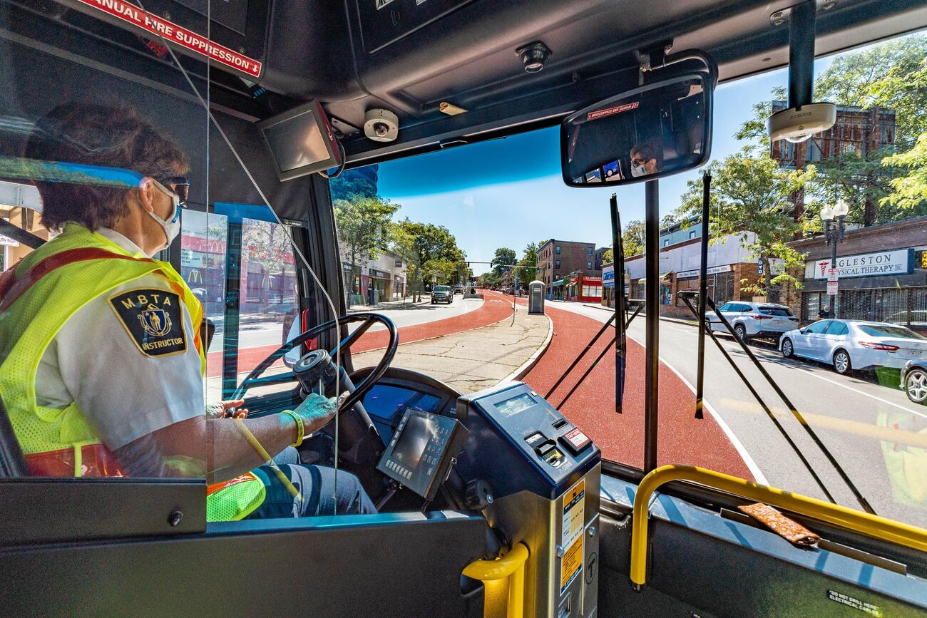 Photo taken from inside a bus of a masked driver with an "MBTA instructor" shirt patch driving down the columbus ave center lane