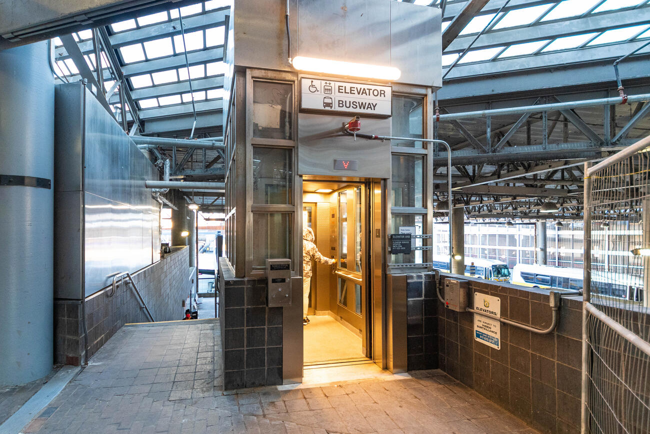 Elevator 848 is open and lit from inside, and a passenger is inside at the far end of the pass-through elevator, ready to be taken down to the lower busway. Stairs down are visible at right, and the busway can be seen at right.