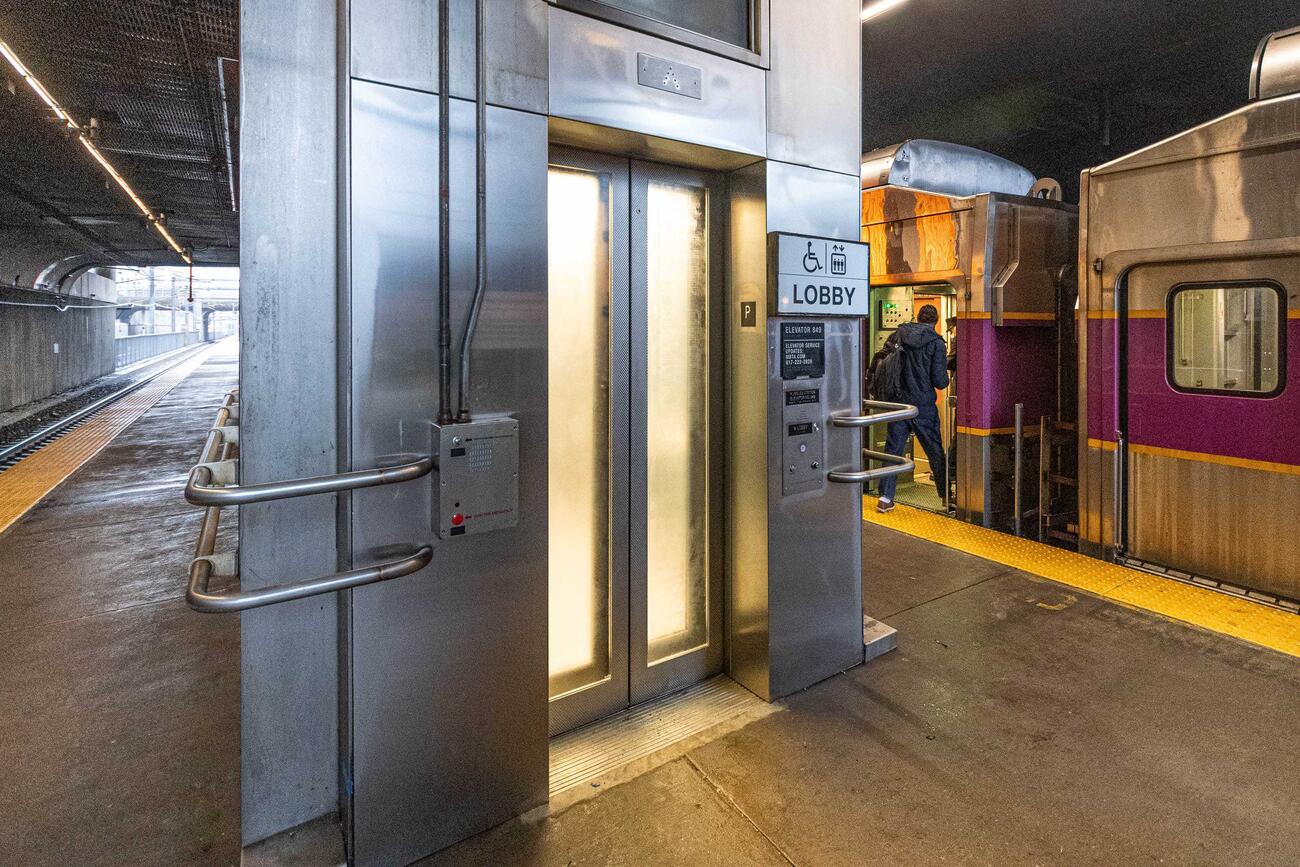 The newly reopened elevator 849 is shown closed, light streaming through its translucent doors. The elevator is on the center of the Commuter Rail platform at Ruggles station. A passenger can be seen boarding a Commuter Rail train at the right side of the platform.