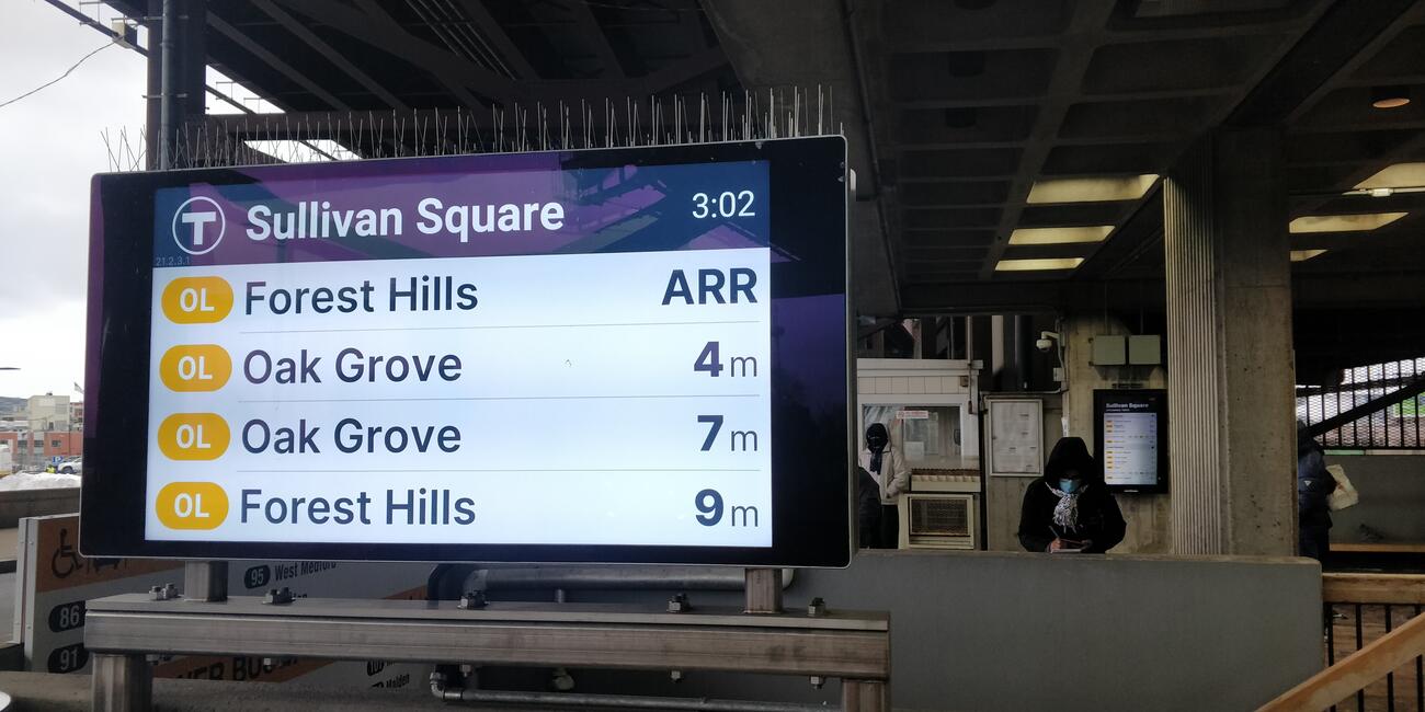 At left is a close up of a digital sign displaying Orange Line arrivals; at right are masked riders. Sign reads as follows: Sullivan Square and the time (3:02) are on the top line, and the following four lines say Forest Hills ARR, Oak Grove 4 m, Oak Grove 7 m, Forest Hills 9 m.