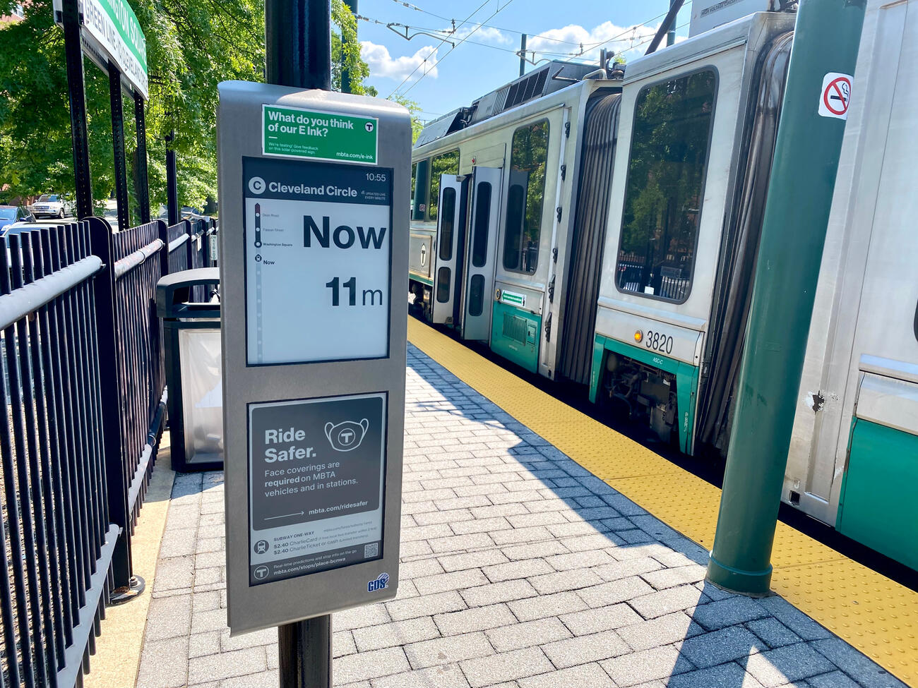 At left is an E Ink solar-powered digital sign at the Washington Square trolley stop on the Green Line's C Branch. The E Ink sign shows the time (10:55) and the destination (Cleveland Circle), and says Now in large letters to show that the trolley is now boarding. Below "Now" the sign says "11 m" to show that the next trolley will arrive in 11 minutes. A Green Line train passes on the tracks at right. 