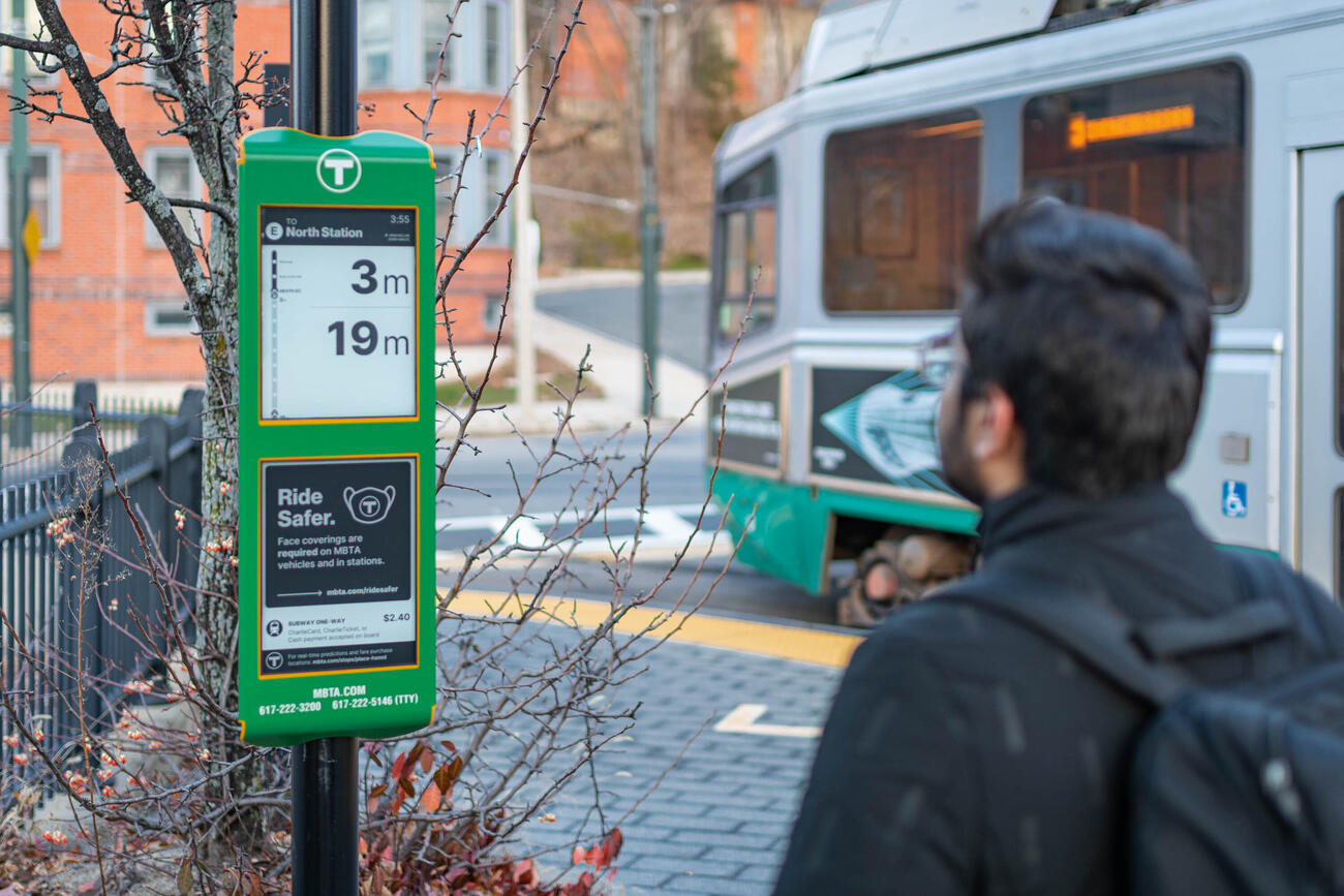 A rider reads predicted arrival times on an E Ink digital sign at the Heath Street trolley stop on the Green Line's E Branch. The E Ink sign has a green border to match the related trolley line.