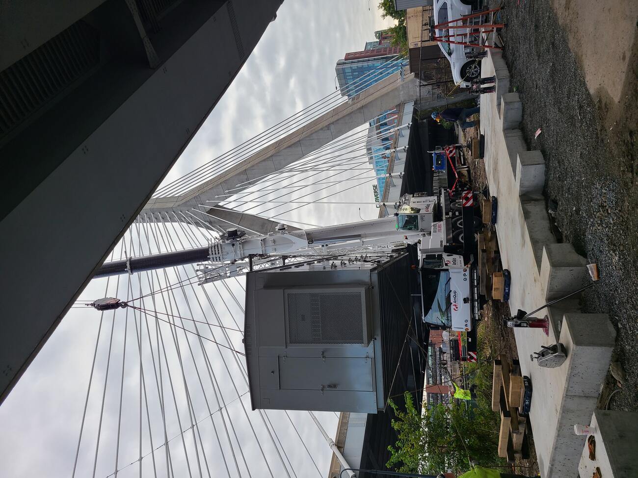 A grey metal building is suspended by a crane before it is set down on a poured concrete pad by the Zakim Bunker Hill Bridge at North Station.