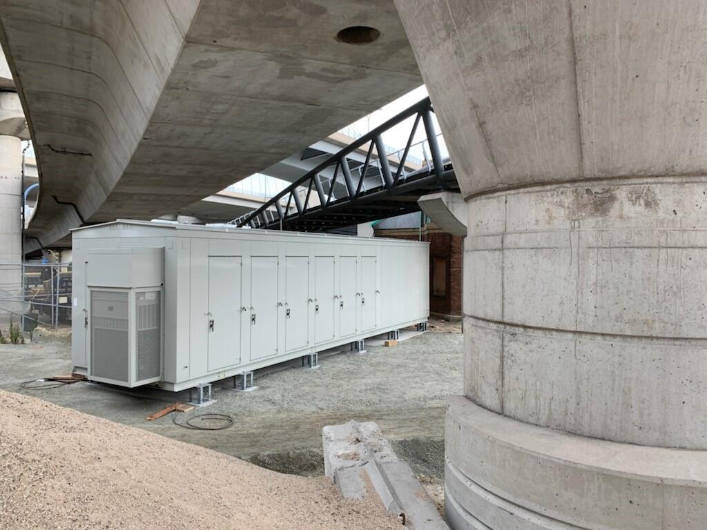 A grey metal trailer used as a signal house sits below below a poured-concrete underpass at North Station—the house shelters Commuter Rail signal cables 