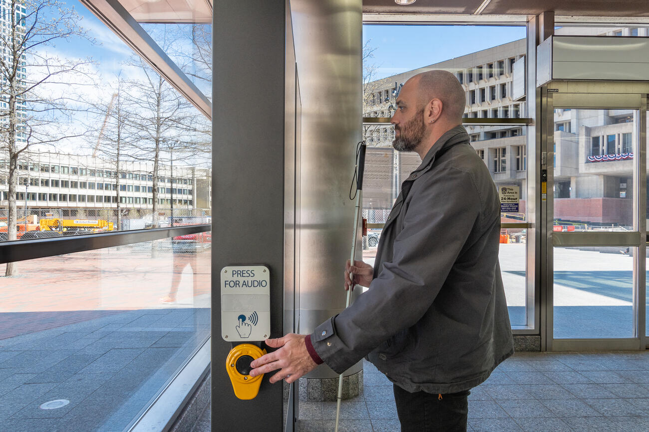 A blind man holding a white cane is seen in profile standing before a digital sign. His left hand is on a button labeled PRESS FOR AUDIO in standard writing and in braille. The rider stand in the lobby of the Government Center station; through the windows behind him the Government Center buildings are shown at right. 