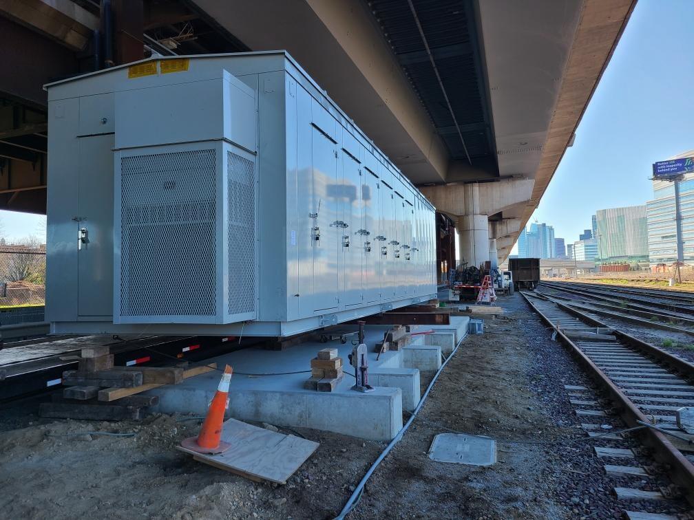 A portable grey metal building with numerous doors sits on a poured cement foundation under an overpass. At right are empty Commuter Rail tracks. tall rectangular buildings ca be seen beyond the tracks, with a blue sky overhead.