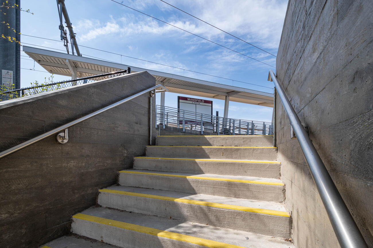 In the foreground are six steps up to the Mansfield station platform. Each step has a bright yellow stripe painted at its edge to improve visibility, and steel bannisters are on the walls that flank the steps. At the top of the steps is a large sign that says MANSFIELD BOSTON on a platform covered by a canopy under a bright blue sky. 