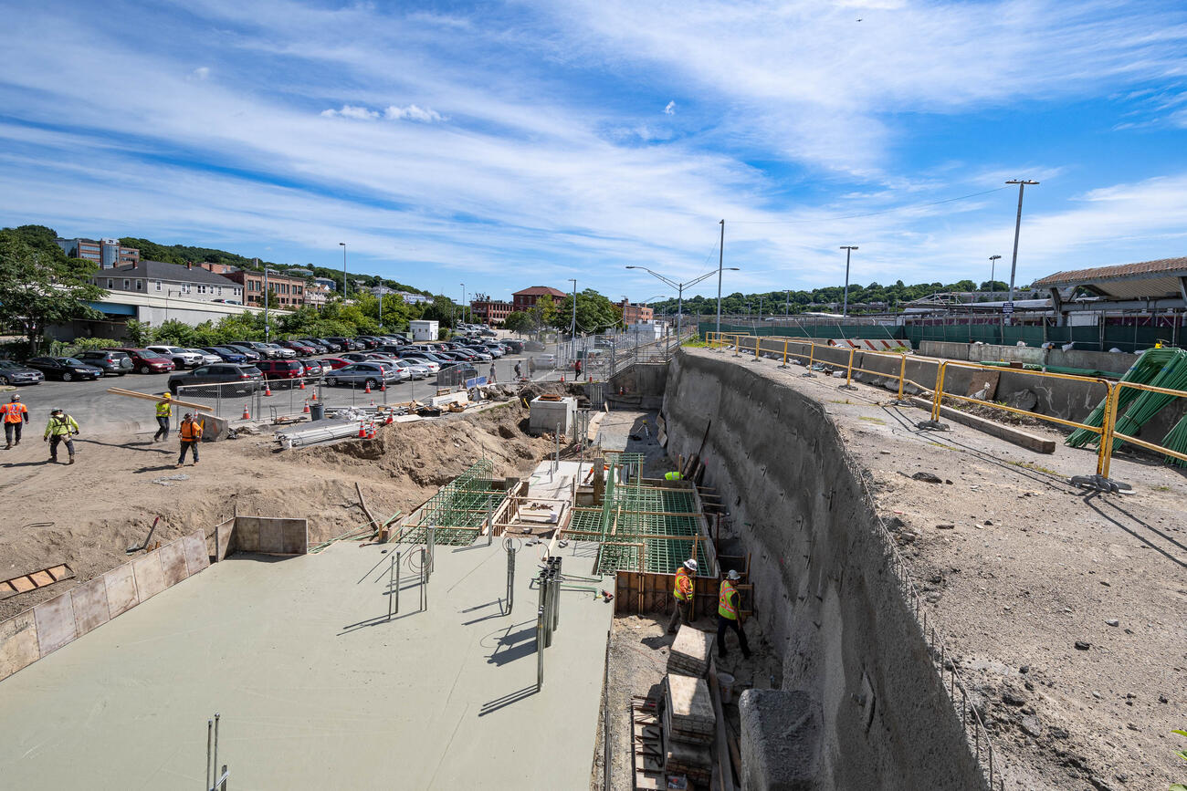 Construction workers in a wide trench next to the station's parking lot install reinforcement and concrete placements for an elevator, retaining wall, and north headhouse foundations.
