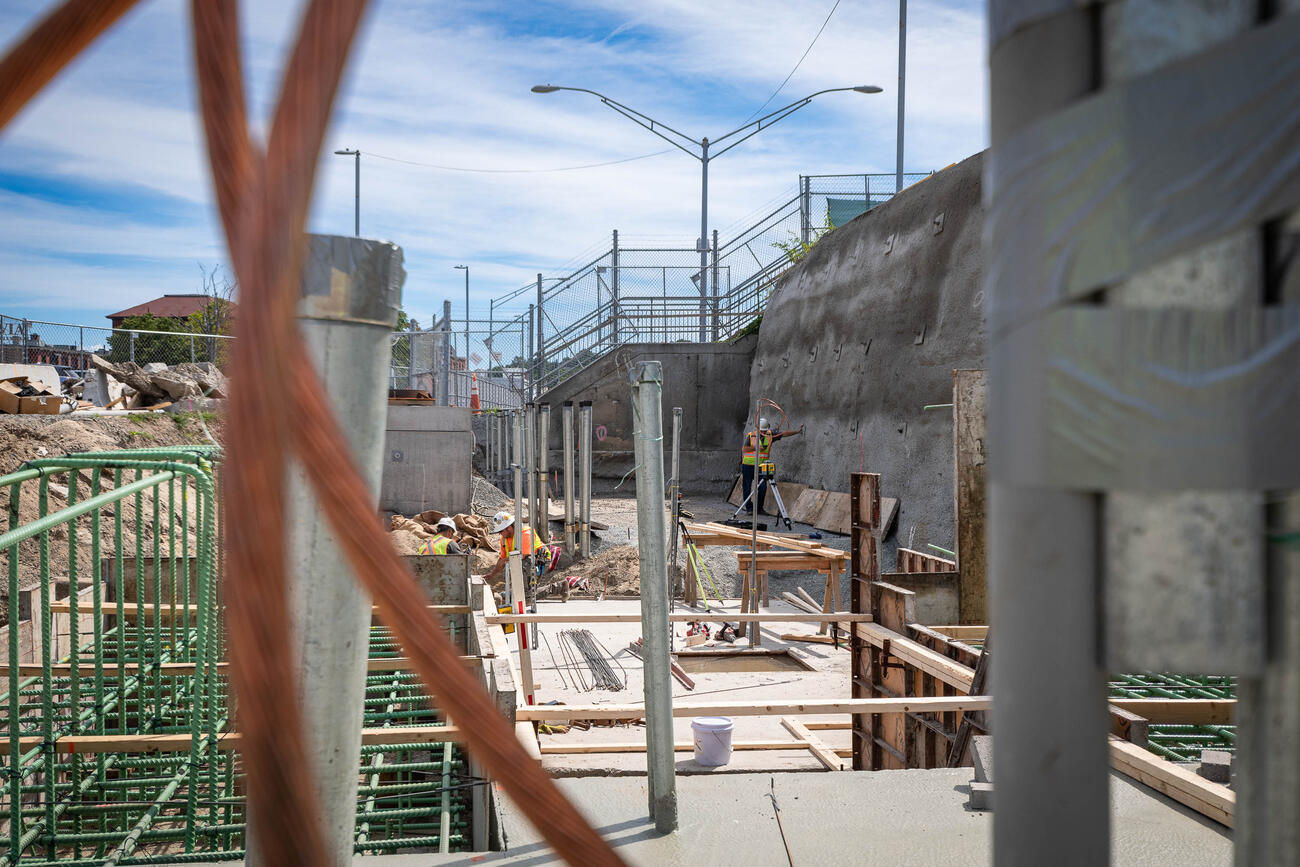 A construction worker in a wide trench next to the station's parking lot installs electric conduits for cables. The cables will power the electrical room, communications room, and an elevator. Blurry copper cables are pictured in the foreground of the construction site.