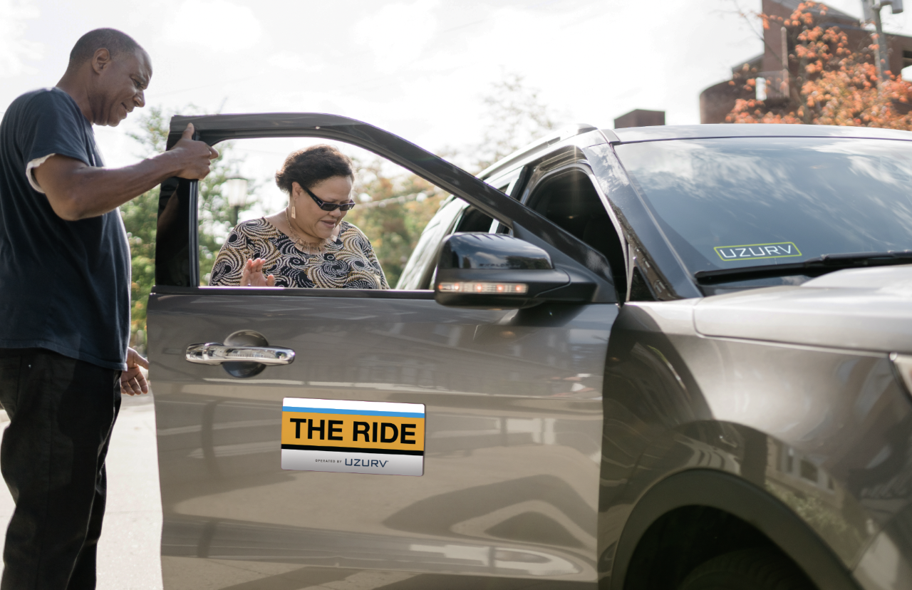 A woman is stepping inside the front passenger seat of a car labeled with a magnetic sign that says the ride powered by uzurv. The driver holds the door open for her.