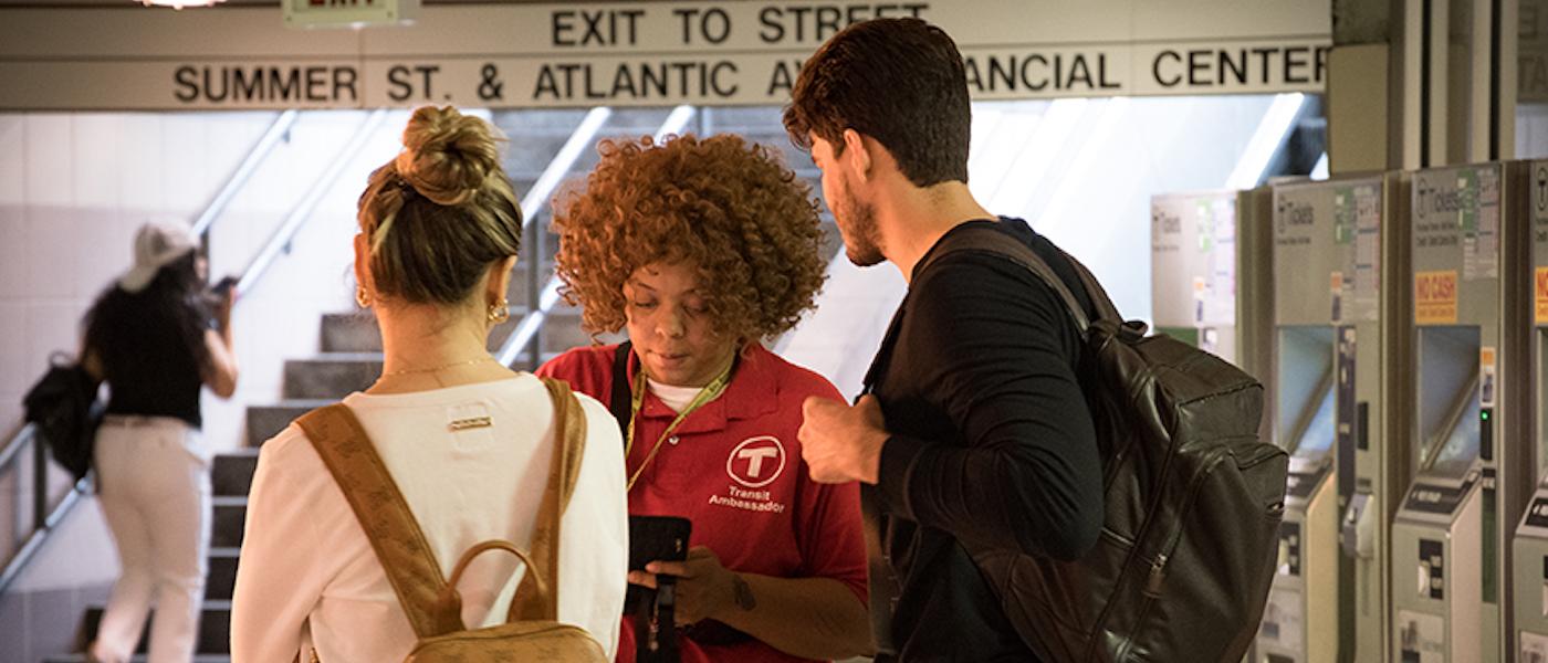 At South Station, a Transit Ambassador assists 2 riders, referencing a tablet.