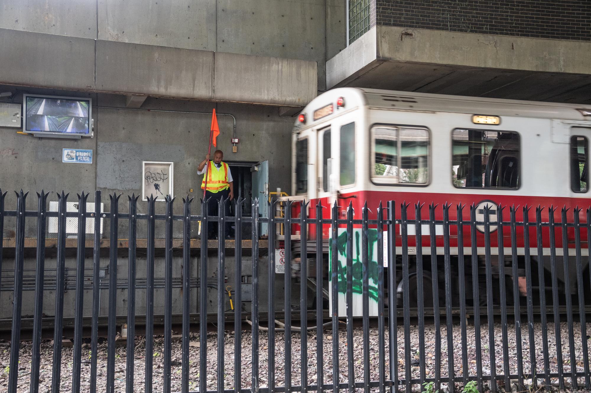 A Red Line train passes through JFK/UMass, with a worker on the platform, holding a flag and acting as a human signal, letting the train know when it's safe to go or when it needs to stop.