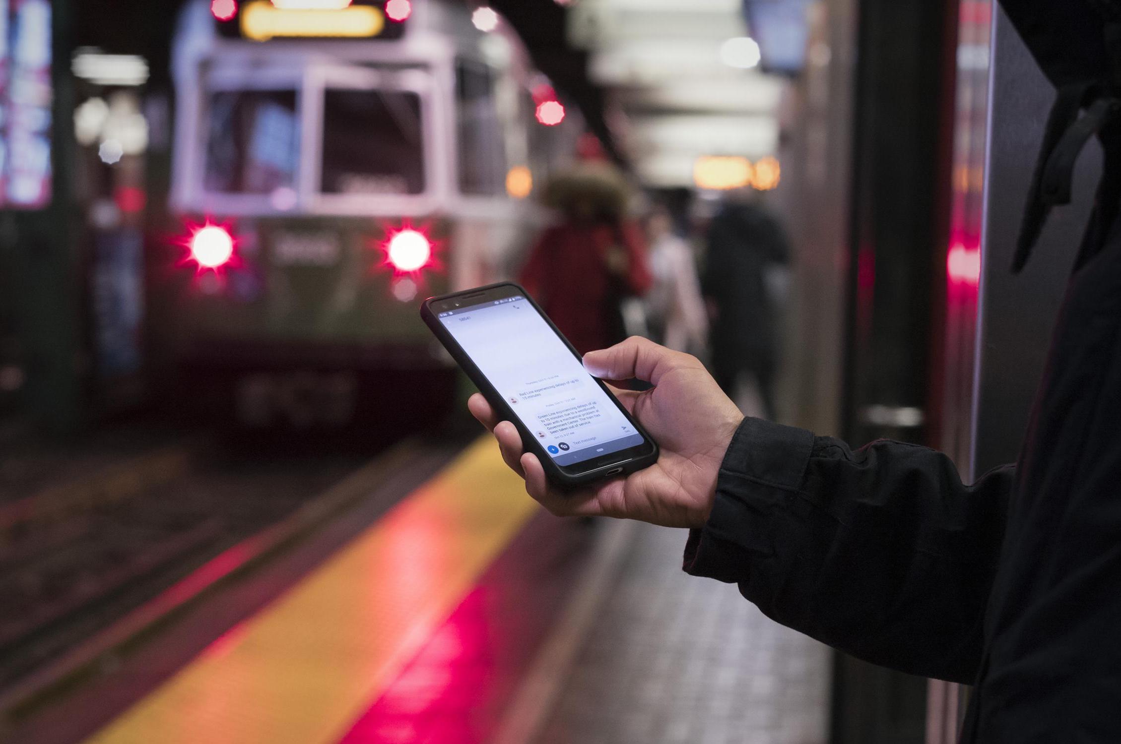 On the platform in Park Street Station, a woman holds her smartphone with a Green Line train in the background.