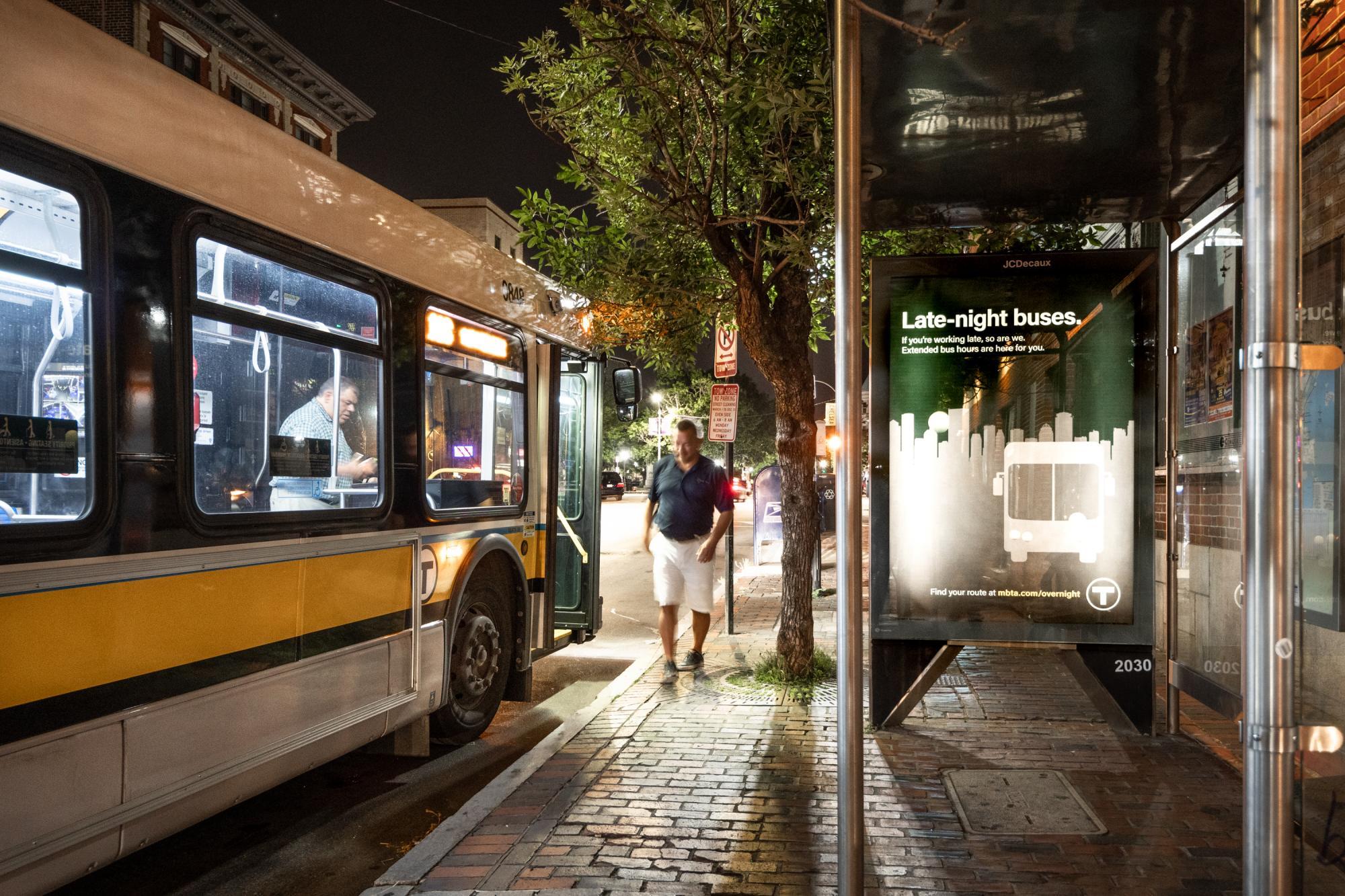 A bus pulled up to a bus shelter late at night, with a rider walking away, and an ad for late night bus service on the shelter wall, which reads,