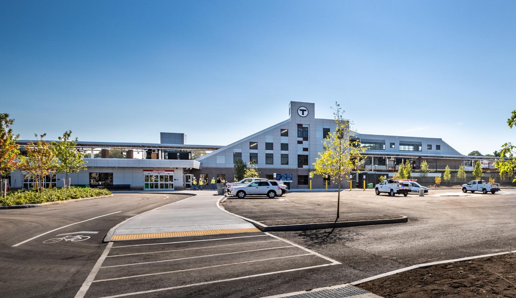 The newly renovated Wollaston Station, viewed from the parking lot, with trees, a bike lane, and sunlight.