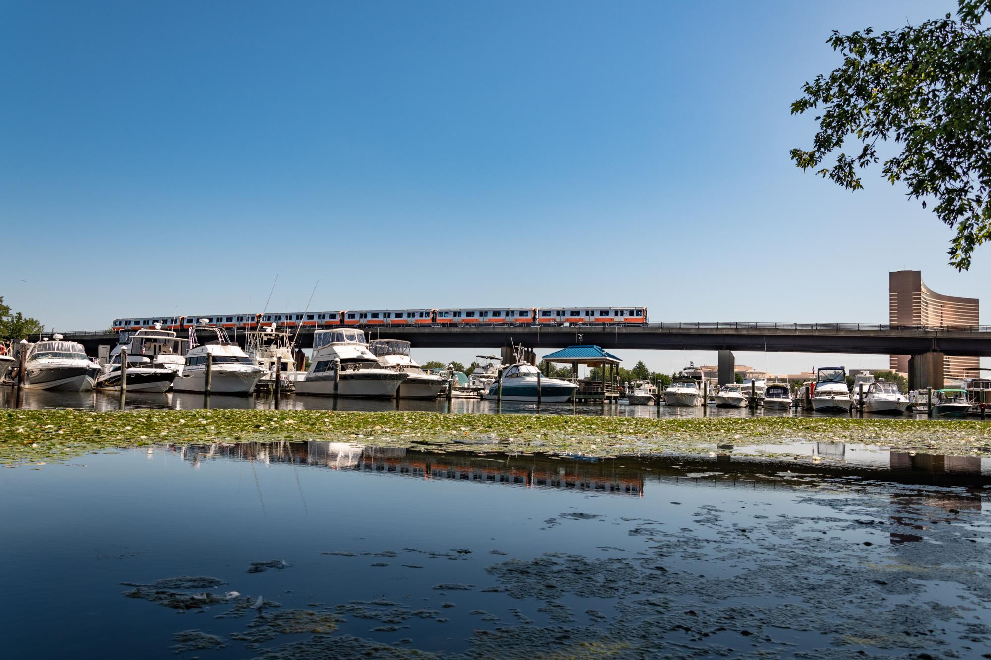 A new Orange Line train travels over the Mystic River, with the Encore casino in the background, boats and water in the foreground, a reflection of the train in the water.