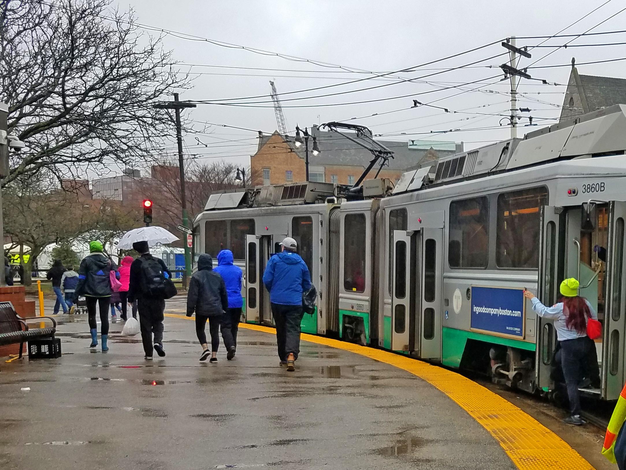 People with jackets and umbrellas walk past a Green Line trolley