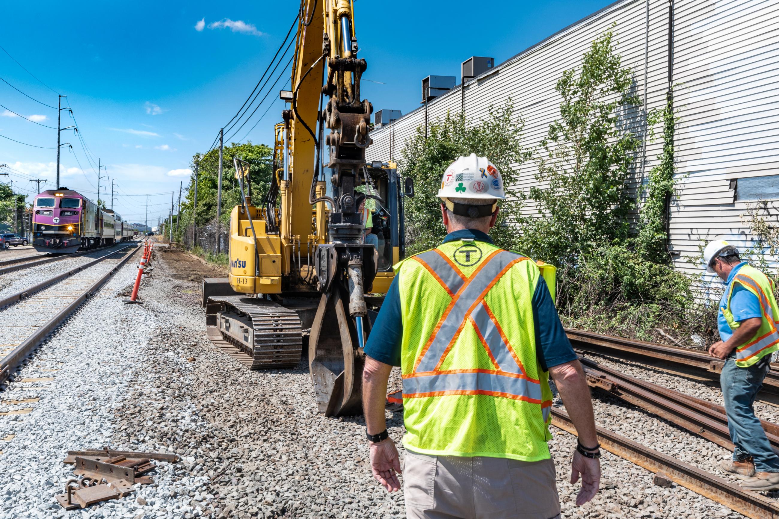 Construction near the new Chelsea Station, with a Commter Rail train coming in on the left, a crew in the middle and and right, with an excavator.