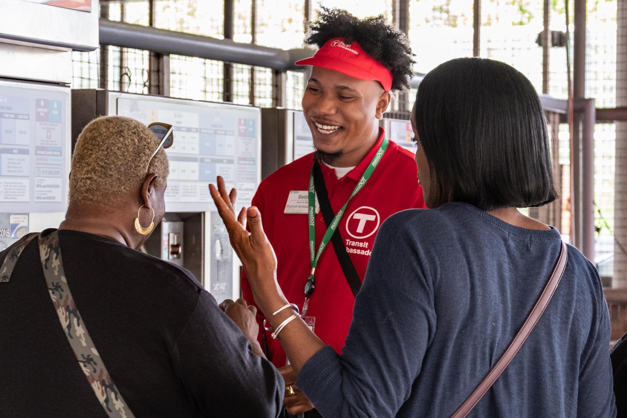 A smiling Transit Ambassador speaks with 2 riders near the fare vending machines at Ruggles Station.