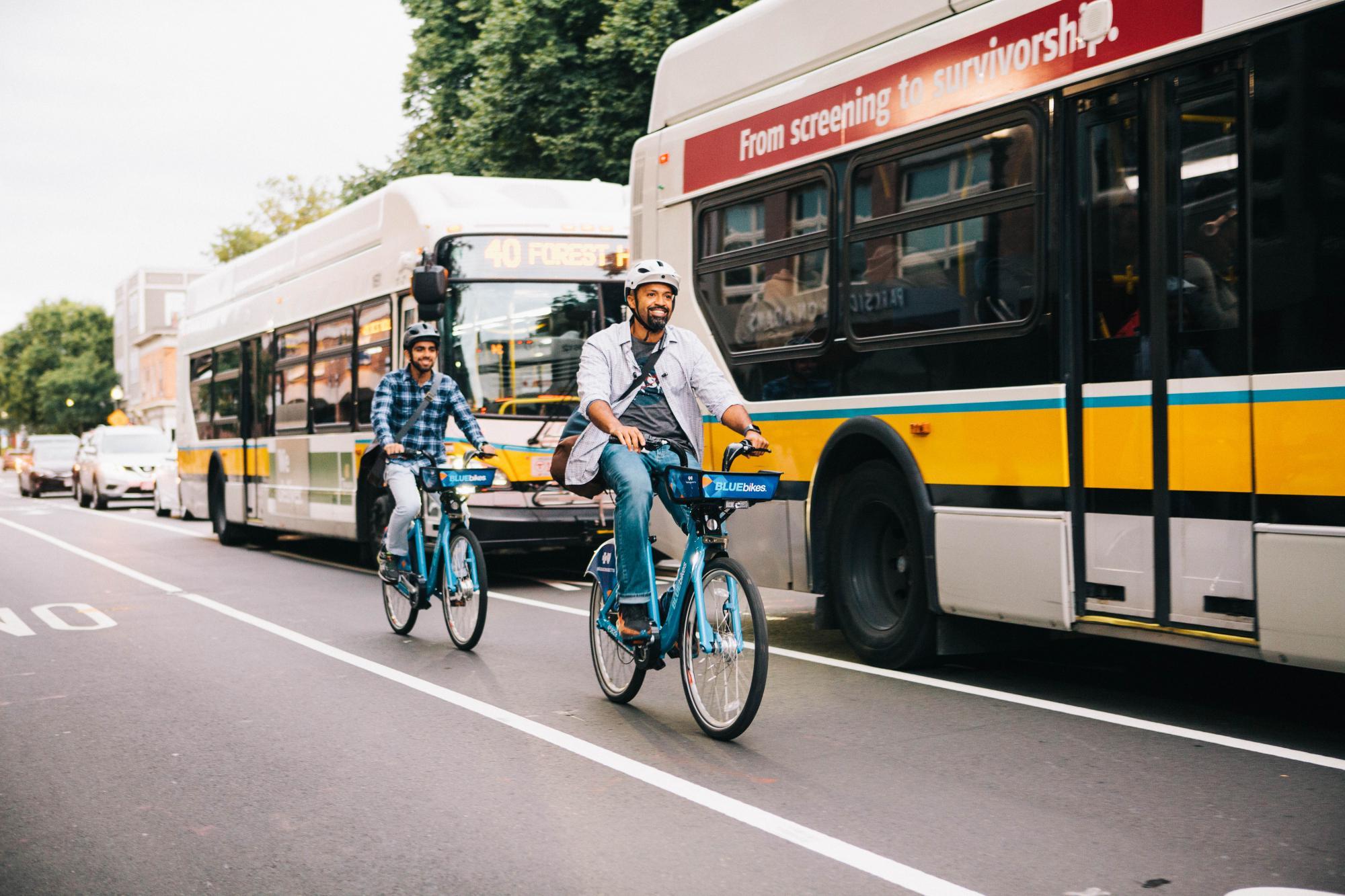Two people ride Bluebikes in the bike lane, with 2 MBTA buses passing by them in the traffic lane. Photo courtesy of Bluebikes.