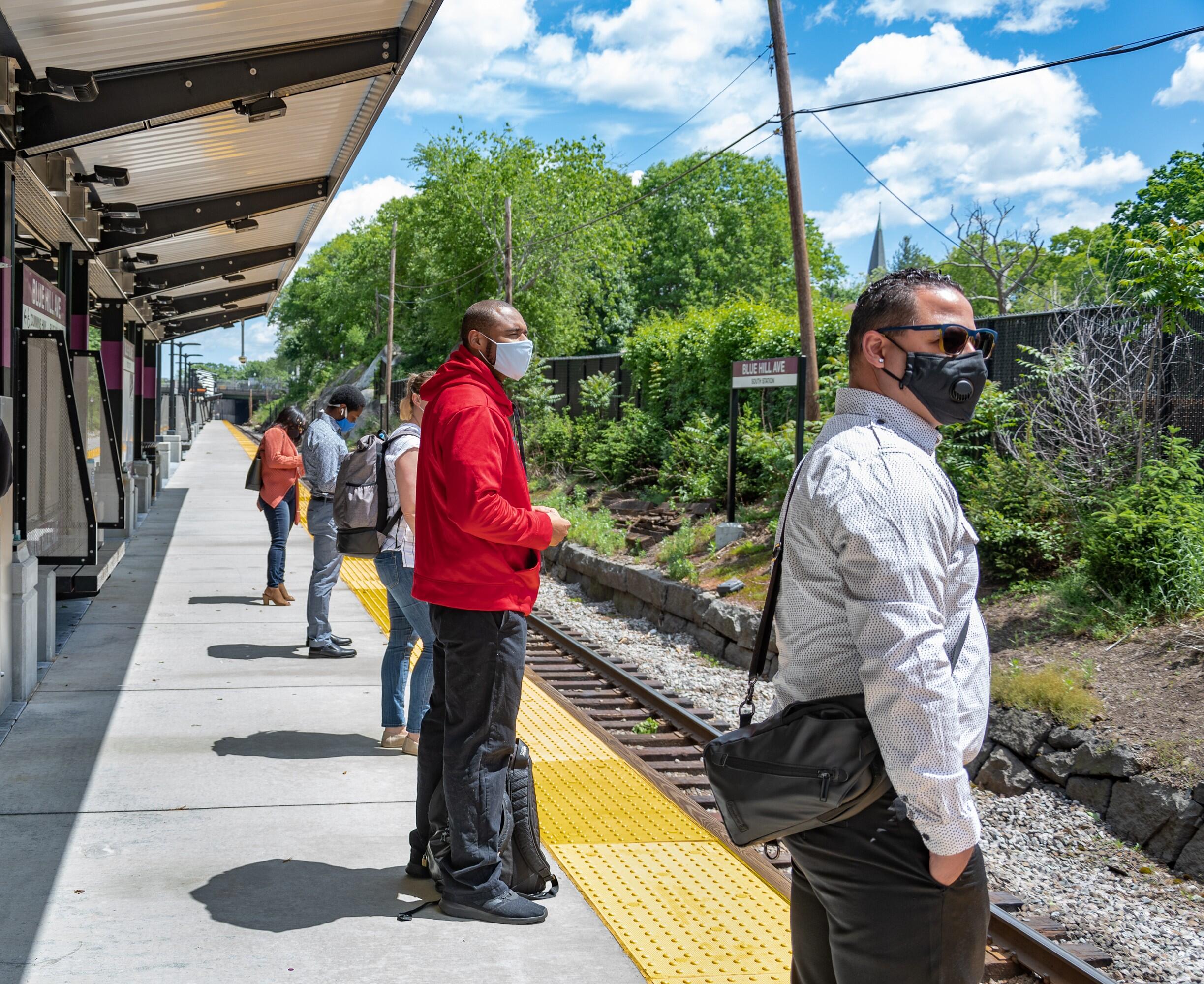 At the Blue Hill Ave Commuter Rail station, riders wearing masks stand on the platform, waiting for the train.