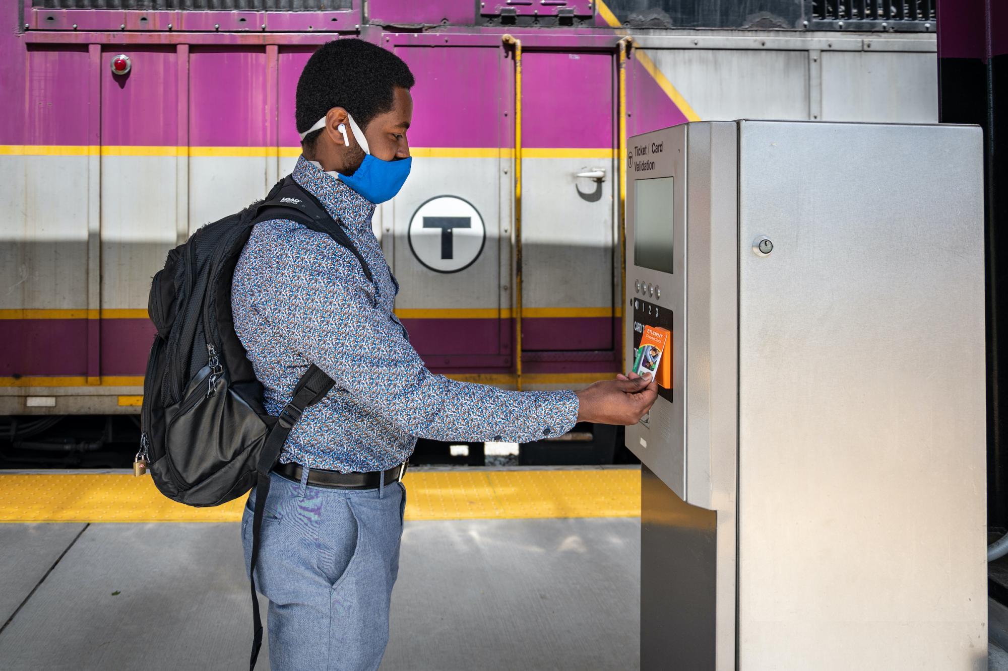 A Commuter Rail rider wearing a face covering uses his CharlieCard at a fare validator at Blue Hill Ave Station.