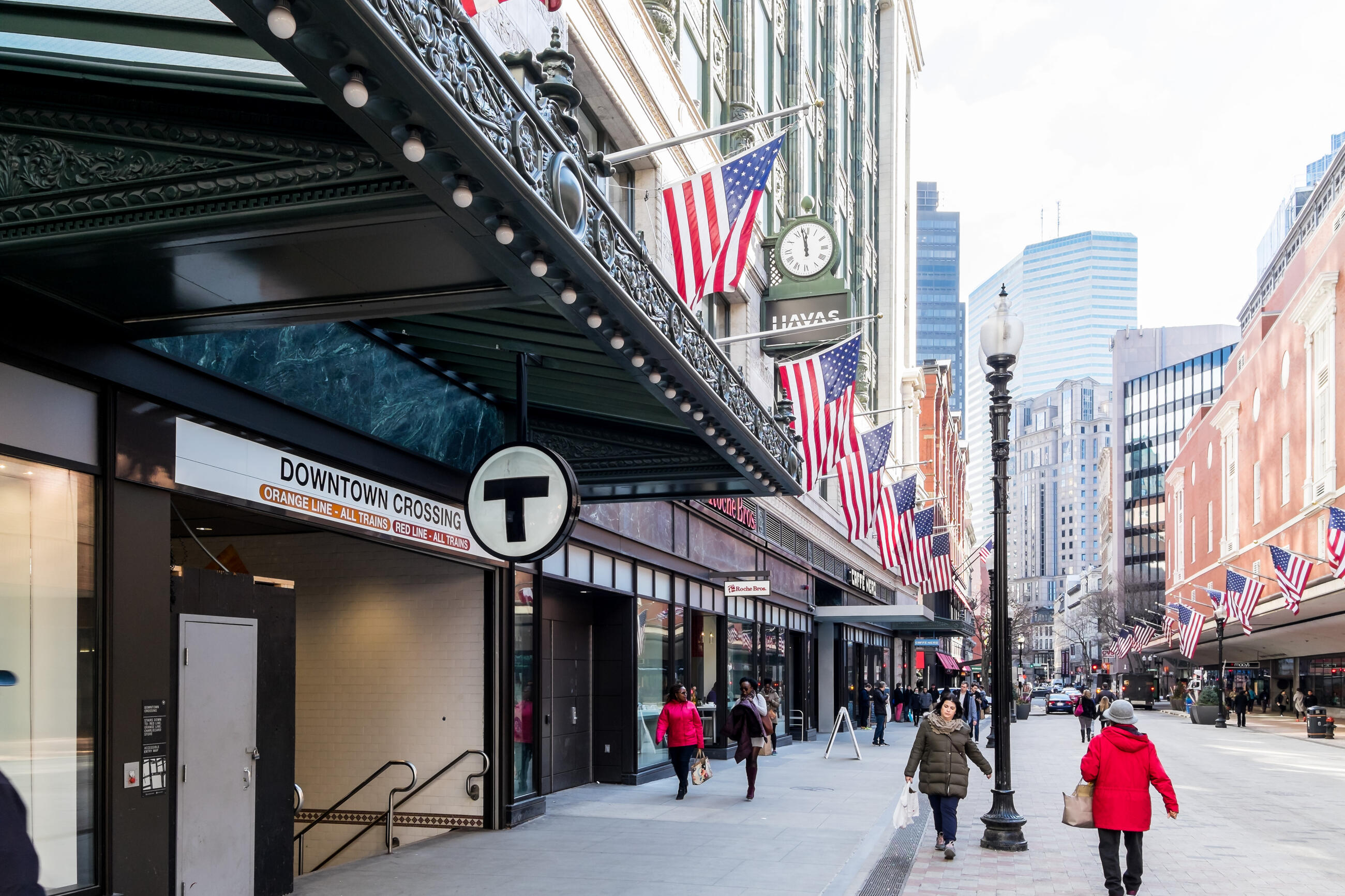 An exterior shot of the Downtown Crossing Station entrance. US Flags line the street, and skyscrapers are visible in the background. Many pedestrians are walking by 