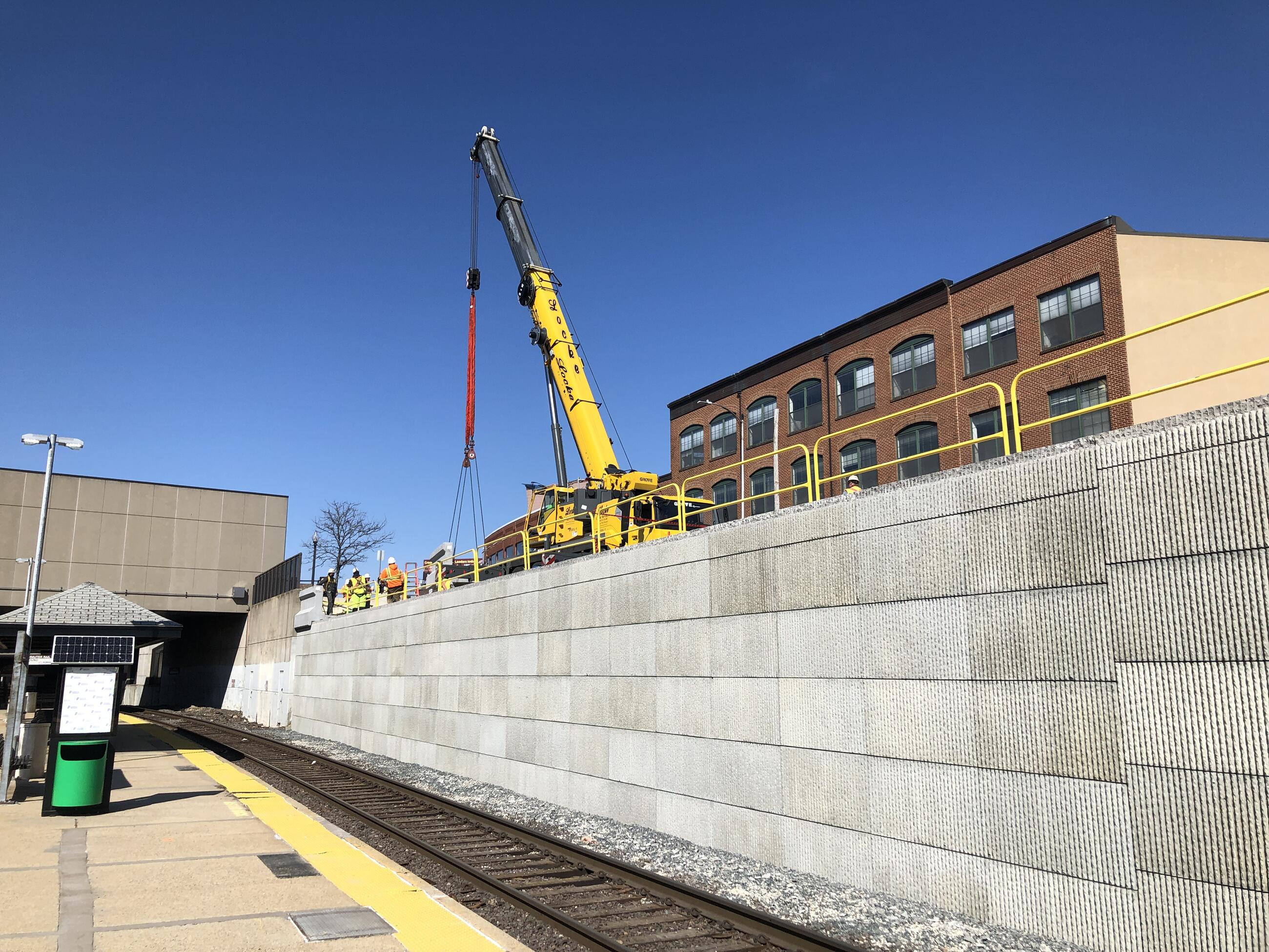 A new concrete retaining wall next to the tracks at Porter Station. Construction workers and equipment are visible on top on the wall.