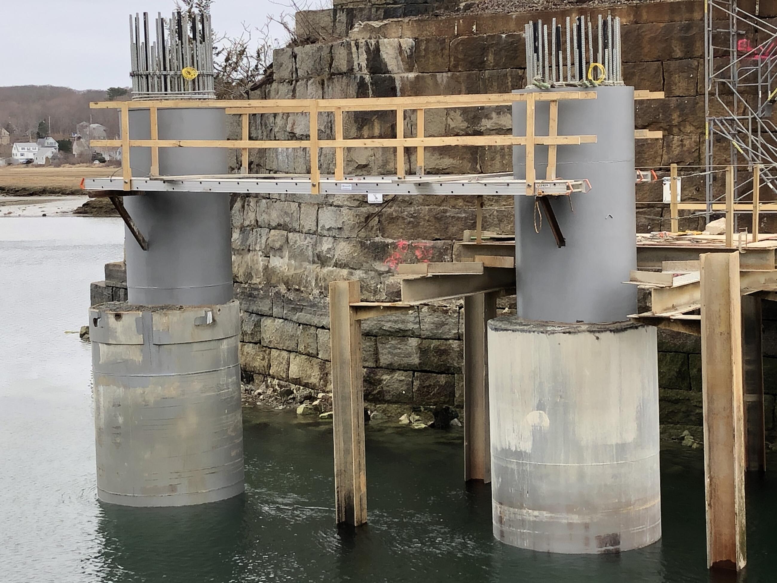 A photo of two large cement shafts in the river with a temporary walkway between them for construction. These will support the bridge above the water