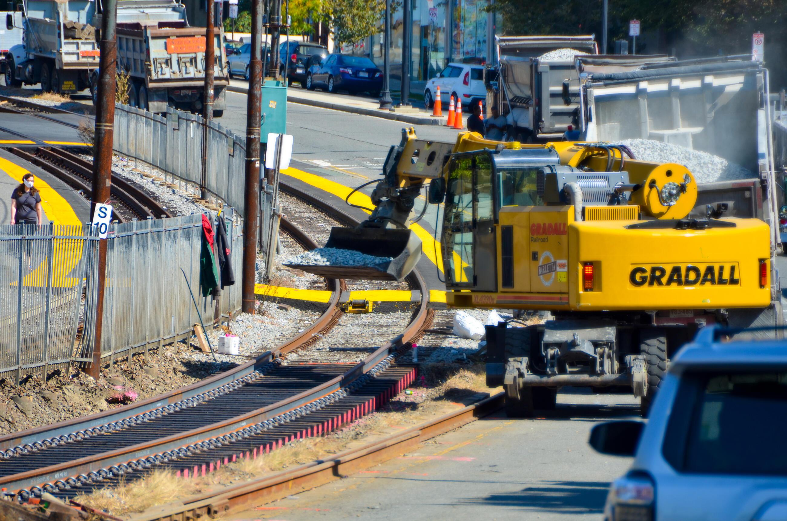 A large construction vehicle moves gravel onto freshly laid track. The track curves around a fence next to a street.
