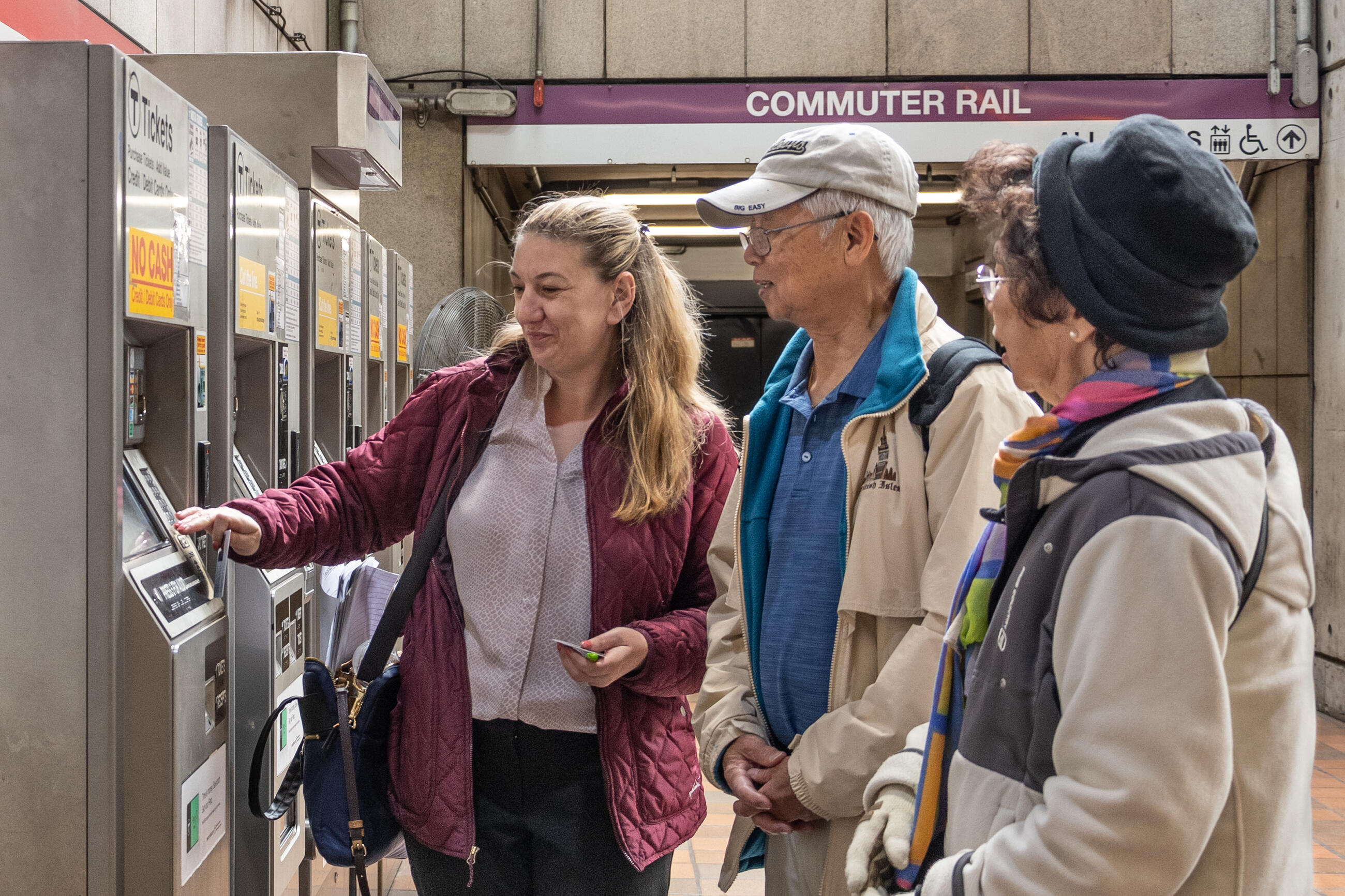 A woman helps two elderly riders purchase tickets at a commuter rail station