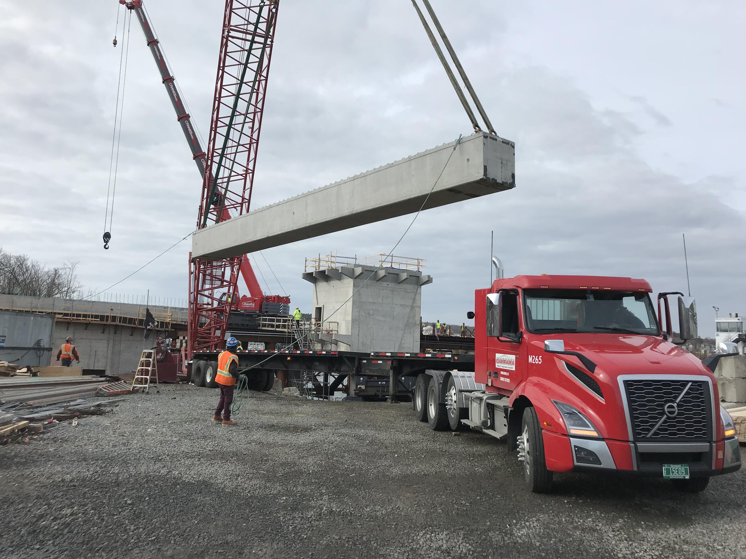 A construction worker in high visibility clothing stands by a semi truck and guides a large concrete support beam suspended by crane 