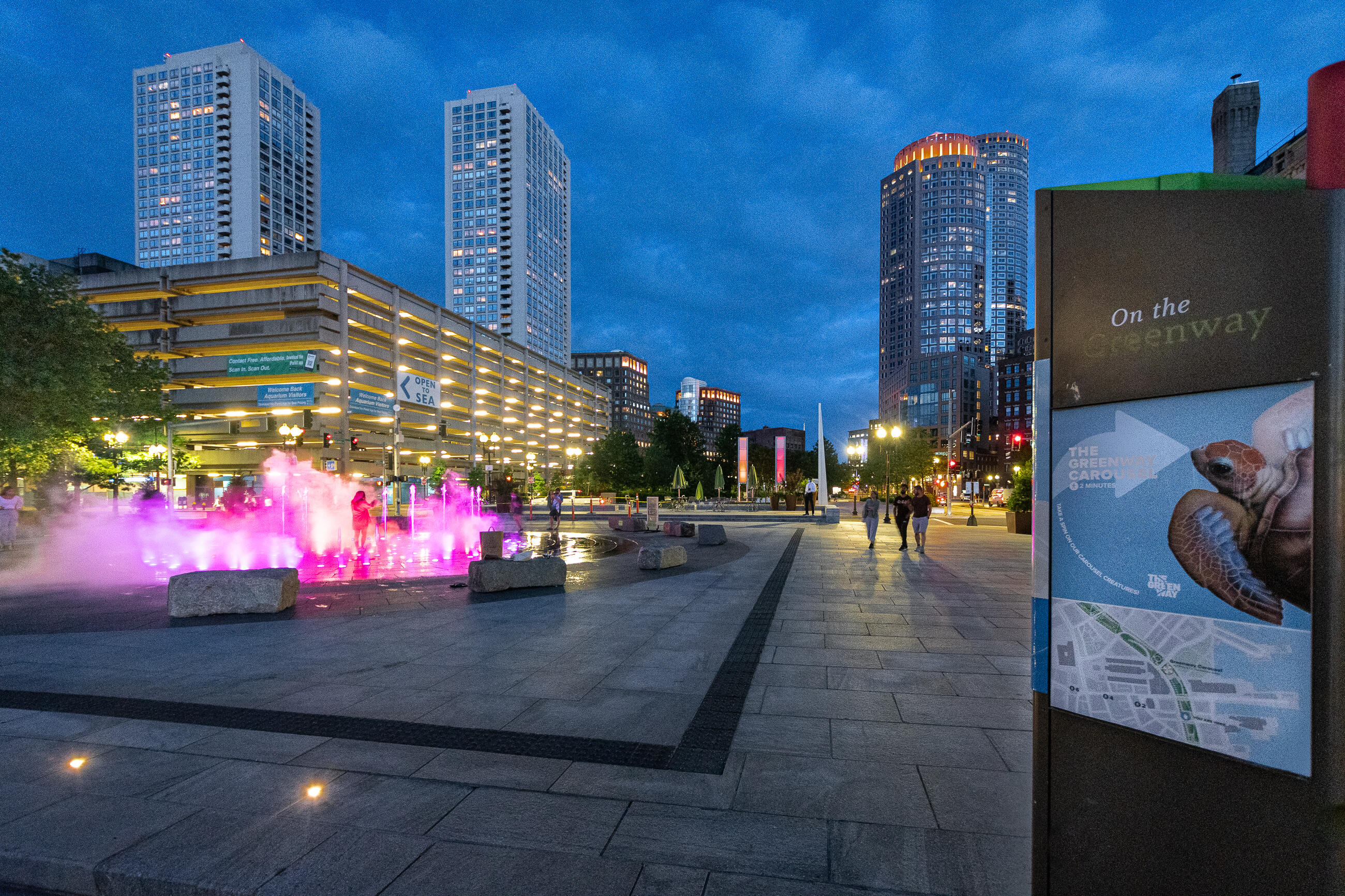 A photo of a big splash fountain at night time, with the Boston Aquarium in the background. It's night time and the fountain is lit up pink and orange. People are taking photos and standing in the pink mist 