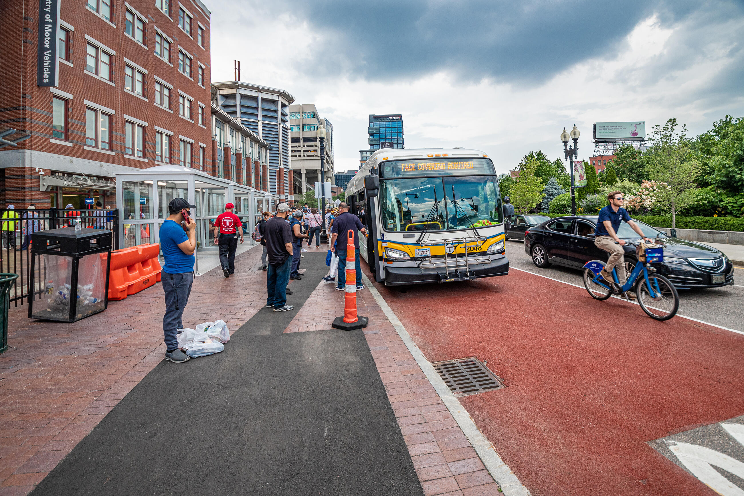 Riders wait in line to board a bus in front of a brick building labeled Registry of Motor Vehicles. The bus, which has a sign spelling out FACE COVERING REQUIRED displayed, is at a Haymarket Station bus stop on Surface Street. Cars and a bicyclist pass to the right of the bus.