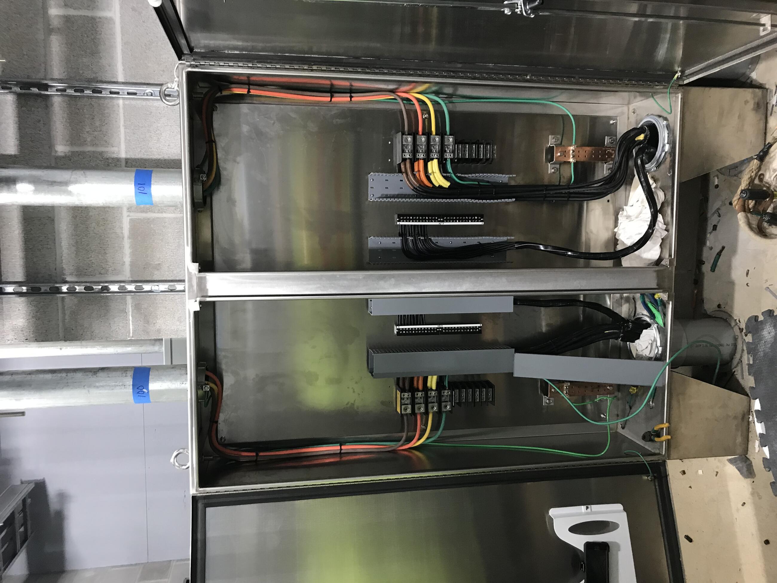 A steel cabinet with two vertical bays has both doors open to reveal thin green, yellow, orange, and brown cables, as well as several thicker black cables. The cabinet is backed by cinderblock walls and is surmounted by two metal ducts into which the colored cables are threaded. 
