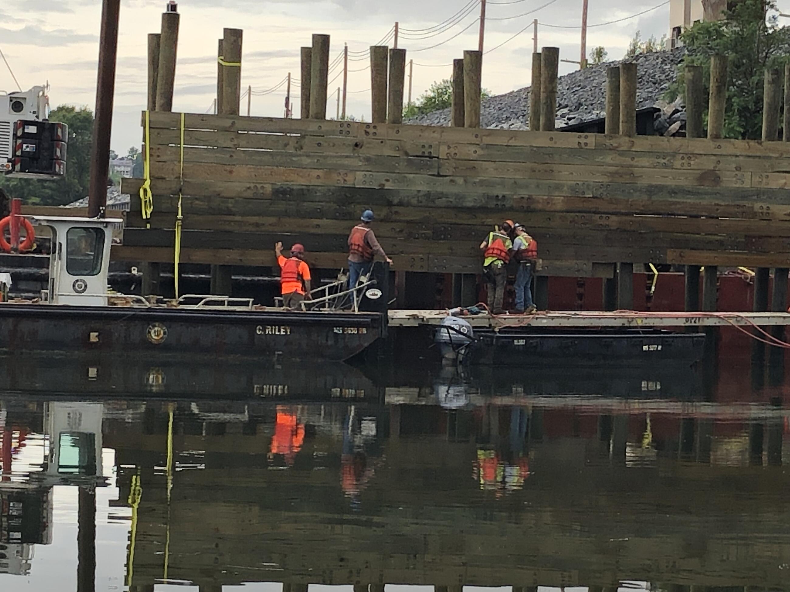 Four workers, three on a platform and one on a boat, install timber boards and beams horizontally to connect tall, round, wooden piers.