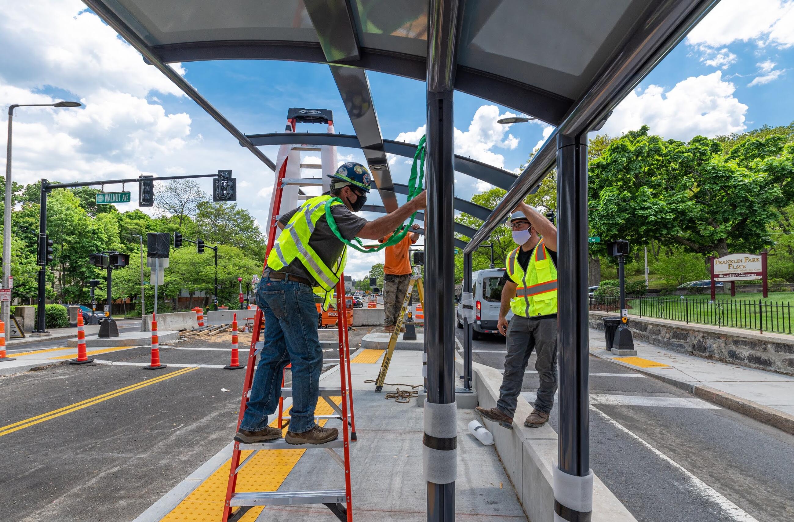 Construction workers in high visibility clothing and hard hats work together to assemble a glass and metal canopy for shelter at the new bus stop