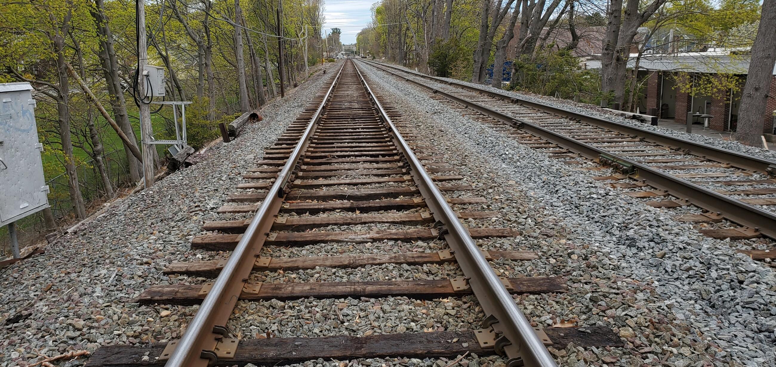 A length of empty Commuter Rail rail and tracks is shown from a vantage point next to the center of the tracks, between the rails, with the track disappearing into the distance at the top. Another empty track is to the right. The two tracks are flanked by gravel and trees, with grass at the left and a building at the right.