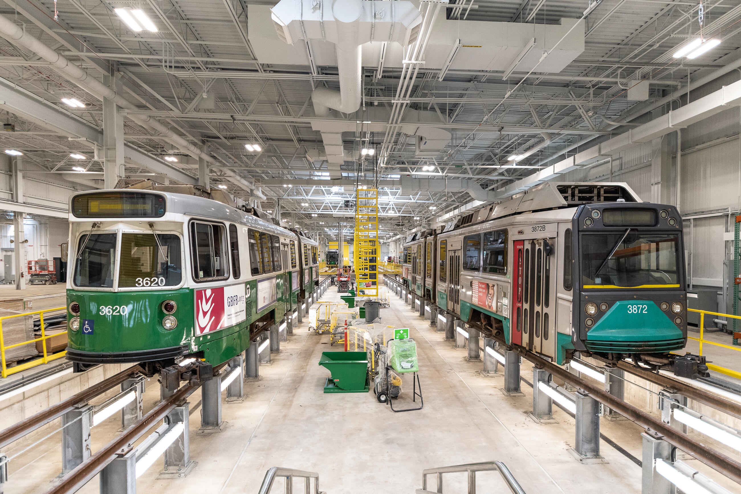 A photo of green line trolley cars at a GLX testing facility. The train on the left is new, and the one on the right is old. They are both elevated on rails inside of a brightly lit warehouse 