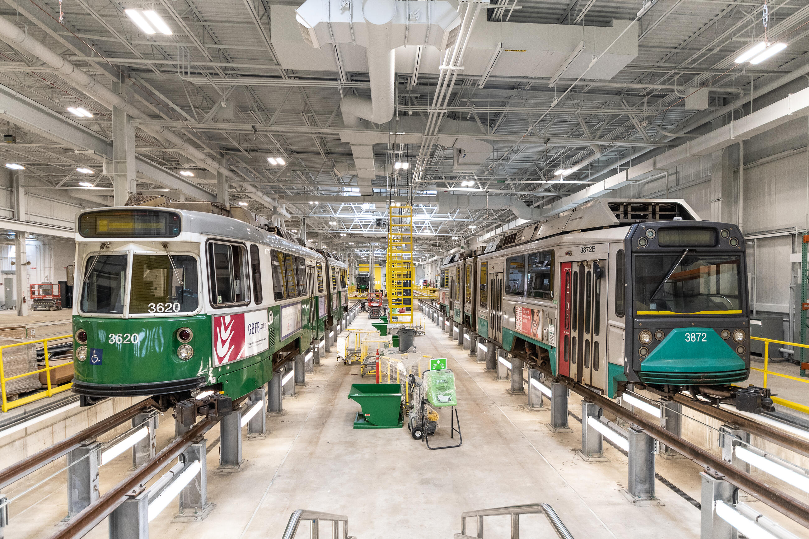 A photo of green line trolley cars at a GLX testing facility. The train on the left is new, and the one on the right is old. They are both elevated on rails inside of a brightly lit warehouse