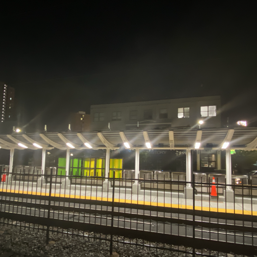 The new Babcock St station is shown at night. No people are visible. The station appears beyond a metal fence, and the new lights are glowing. The new graphic panels have been installed, some bright green, some yellow. Lights show in windows of multistory buildings in the background.