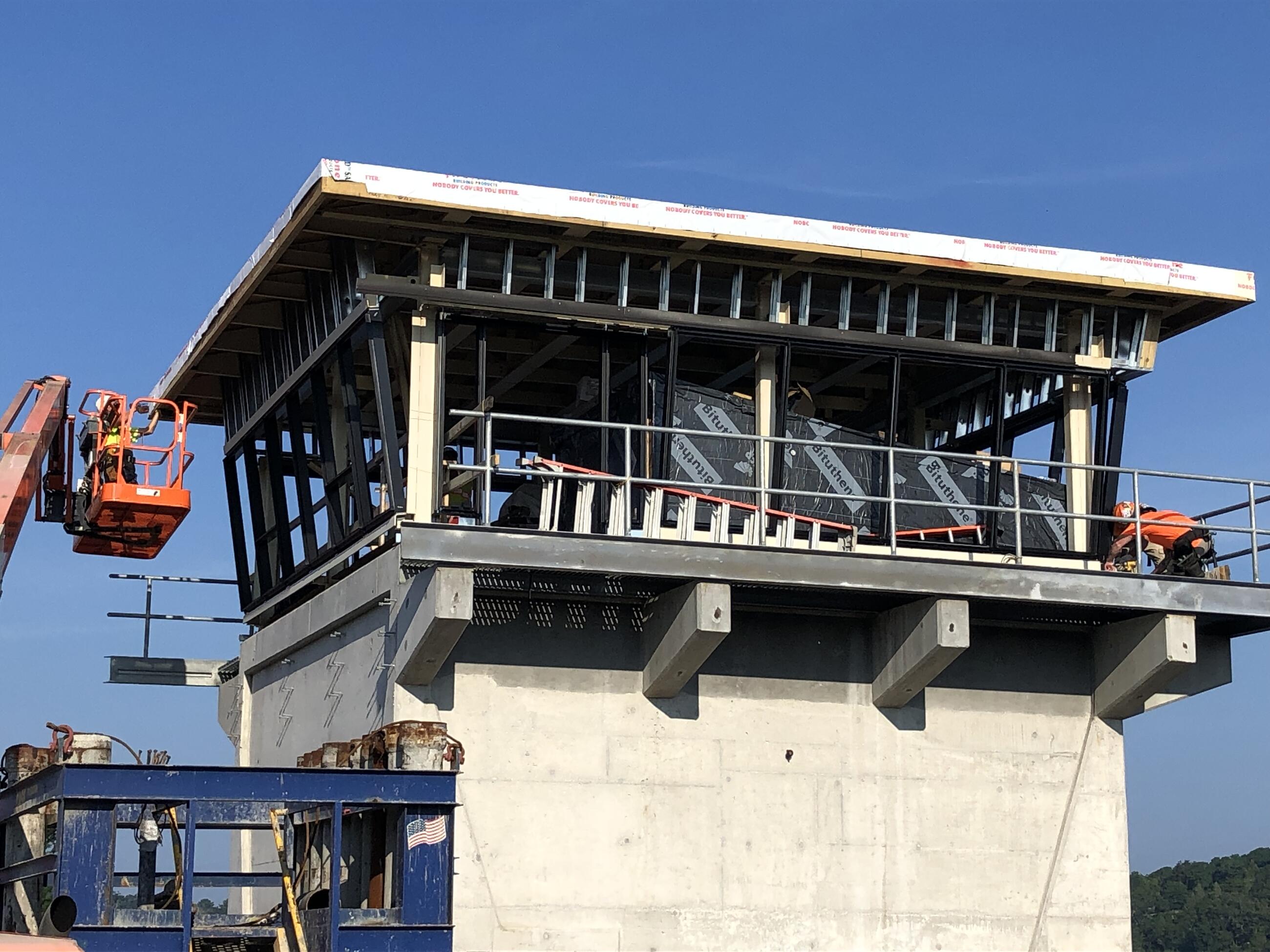 The control tower is shown under construction. A worker kneels on the floor of the observation deck, and another worker is in an orange aerial work platform alongside the observation deck at left. 
