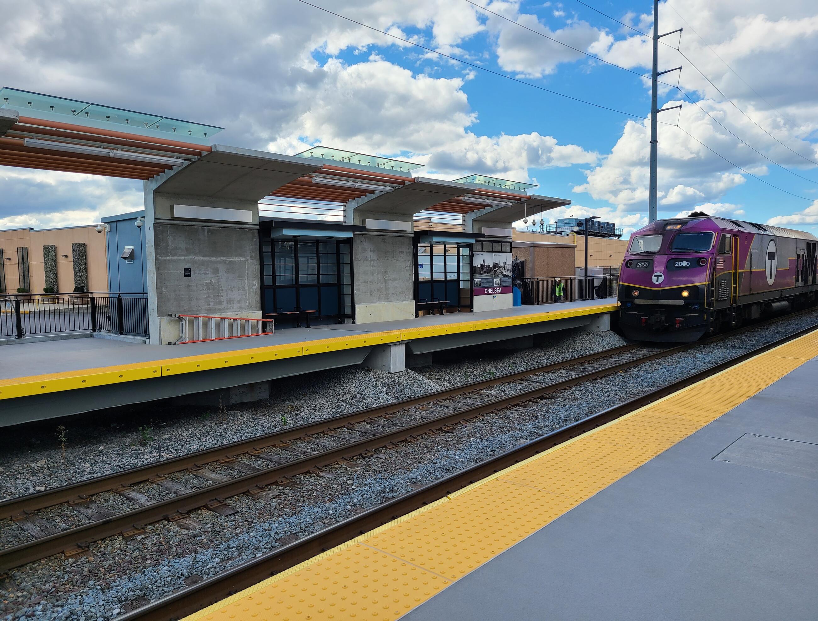 A purple Commuter Rail train is shown at right next to the new station platform. The track runs diagonally from lower left to center right, with the inbound platform at lower right and the outbound platform at upper left. The platform has lighted canopies hanging over outdoor stools in recessed seating areas. A blue sky with white clouds is overhead at upper right.