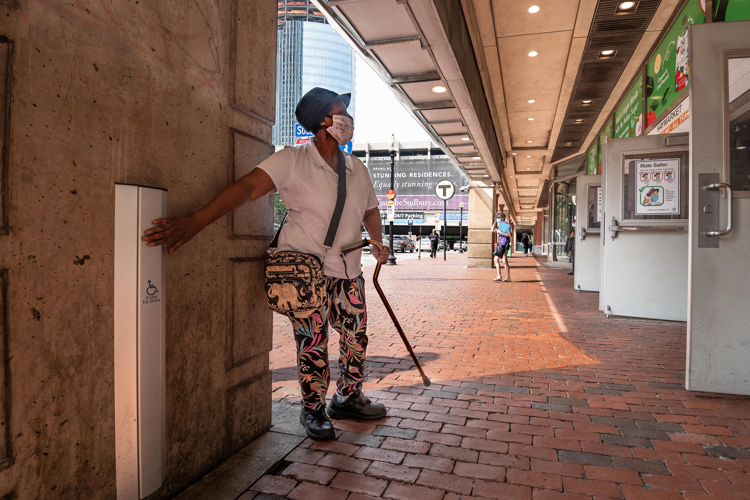 A woman with a cane is pressing the "Push to open" automatic door button and facing the open doors to Haymarket Station